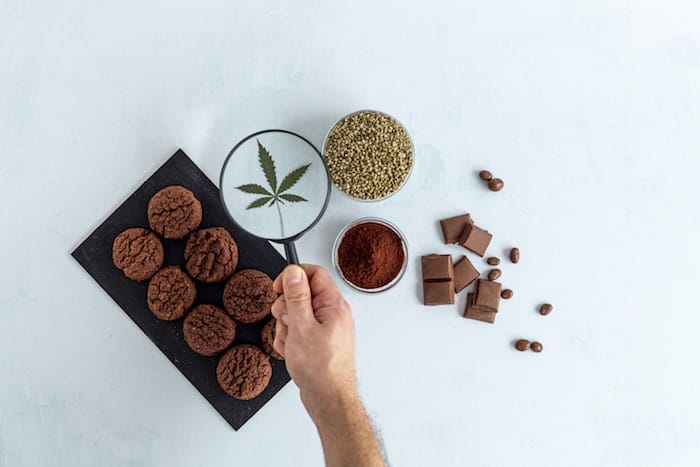 CBD cookies, cannabis chocolate, cocoa and hemp seed on bright background. Hand holding magnifying glass over marijuana leaf. Medicinal marijuana in food.
