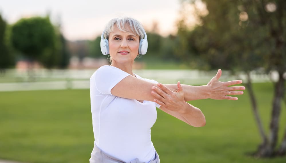 Positive active senior fit woman wearing wireless headphones doing arm stretching, warming up muscles before jogging or outdoor workout, mature female exercising outside in city park in morning