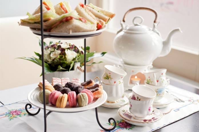 Afternoon tea with desserts and sandwiches organized on a two-tier serving tray beside a white tea pot and several teacups.