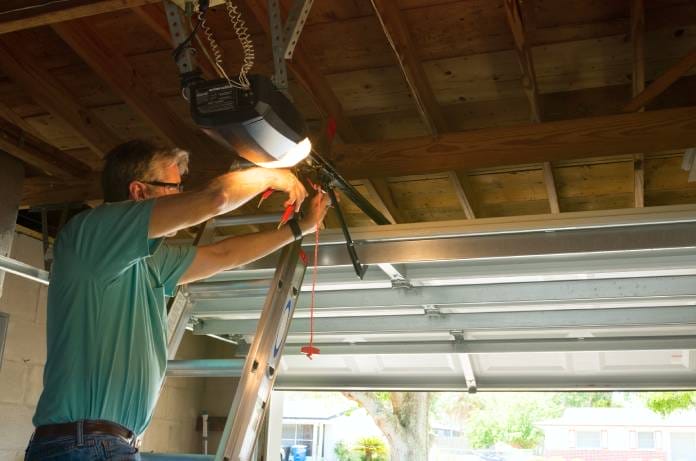 A man in a green polo shirt is repairing a garage door opener. The garage door is open, revealing the yard outside.
