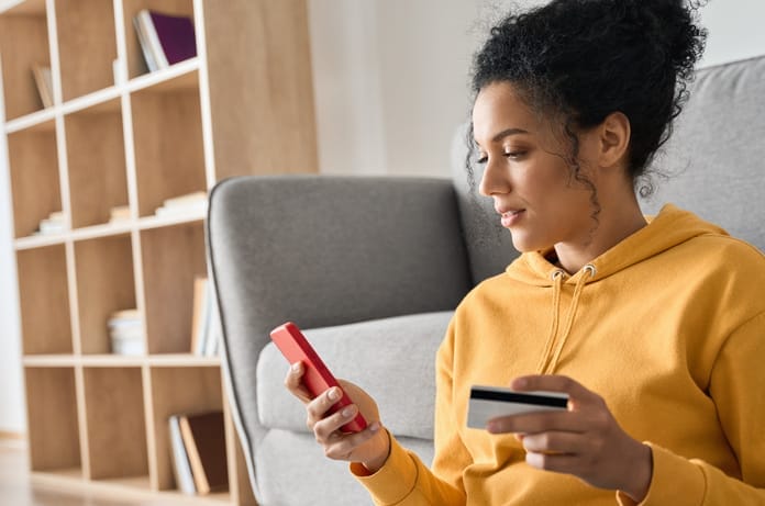 A woman in a yellow sweatshirt sitting on the floor in front of her couch. She is holding a cell phone and a credit card.