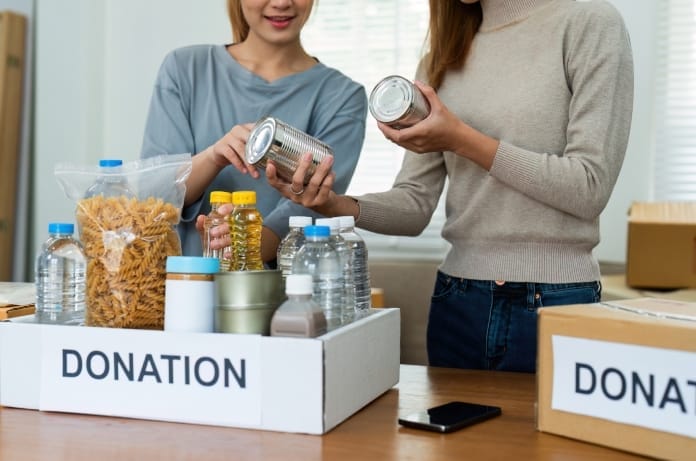 Two women packing donation boxes. They're holding cans above a box with water bottles, pasta, and other food.