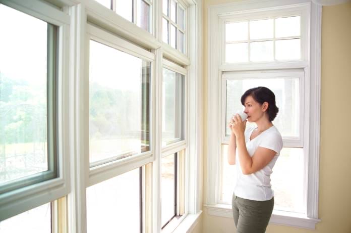 A female homeowner standing next to her windows. She's holding onto a coffee mug in a white tee shirt and pants.