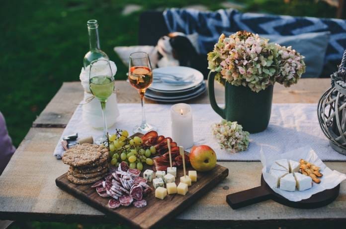 A charcuterie board on an outdoor table with wine glasses and table decorations surrounding the meats, cheeses, and fruits.