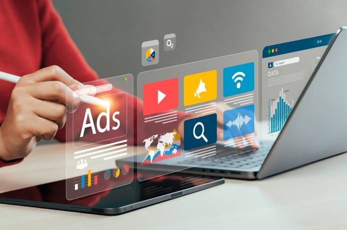 A person in a red shirt sitting in front of a laptop and tablet, using a stylus to interact with digital symbols for advertising.