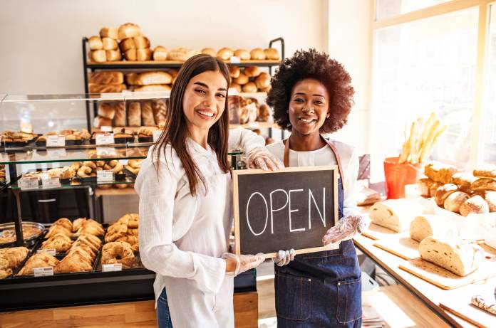 Two bakery business owners standing together in front of their glass display case. They're holding an "open" sign.