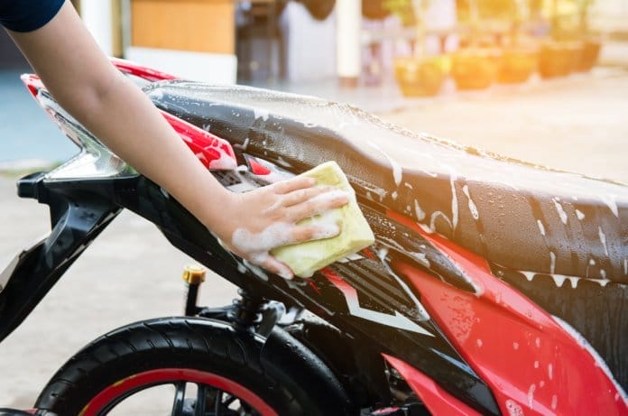 A woman’s hand uses a yellow foam sponge to wash a red and black motorcycle. Soap suds are visible on the bike.