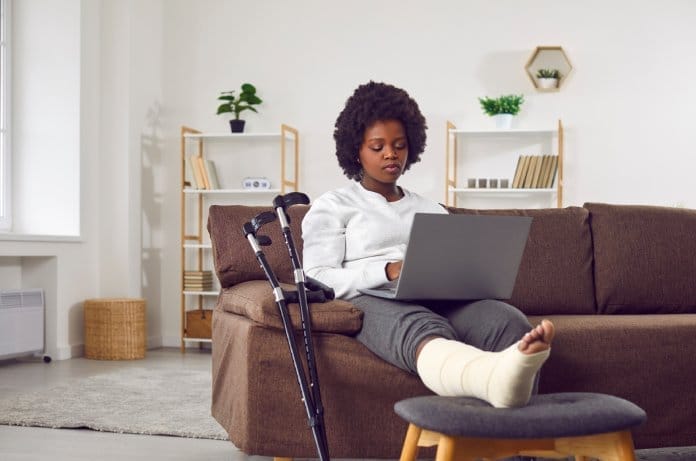 A woman sits on a couch with a cast on her leg. A pair of crutches leans against the couch. The woman is frowning.