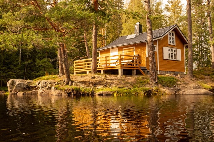 A small yellow cabin with white shutters and a porch sits in the woods surrounded by trees near the edge of a lake.