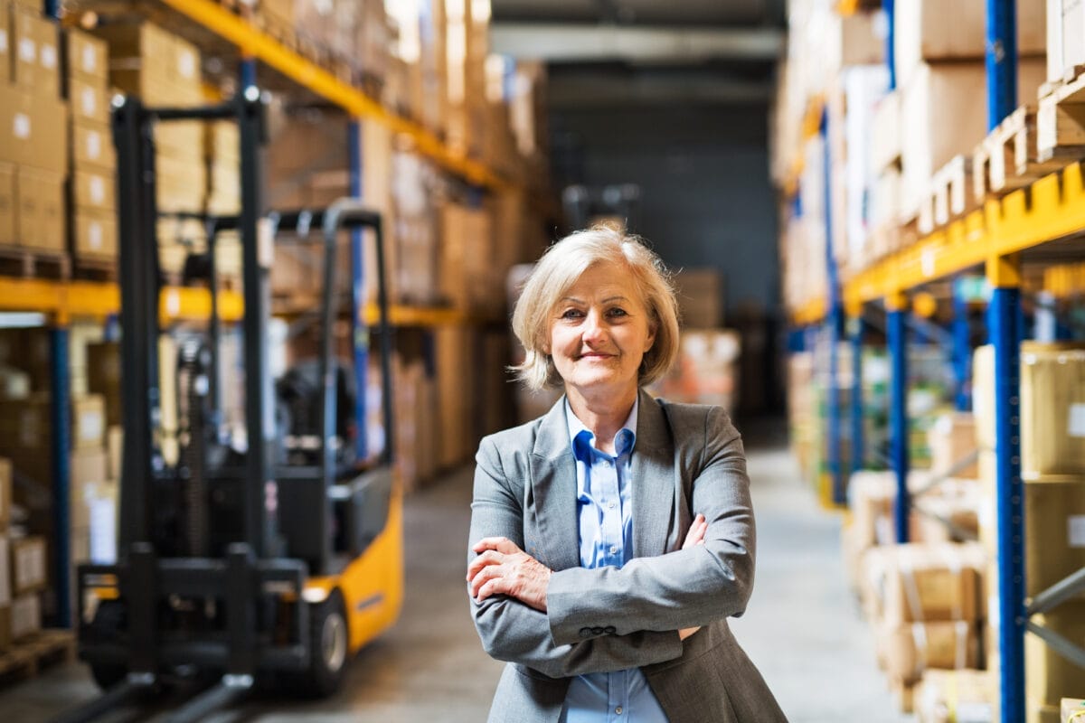 A woman stands in a warehouse with her arms folded. There are shelves on each side of her and a forklift behind her.A woman stands in a warehouse with her arms folded. There are shelves on each side of her and a forklift behind her.