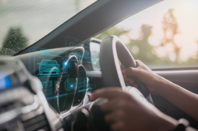 A pair of out-of-focus hands are on a car's steering wheel. The dashboard has graphics of the vehicle and automotive data.