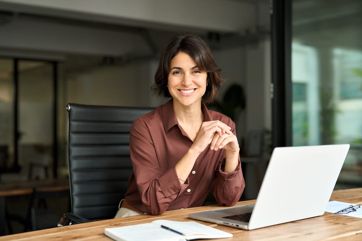 A smiling woman sits at a table with a laptop and an open notebook. She rests her elbows on the table and folds her hands.