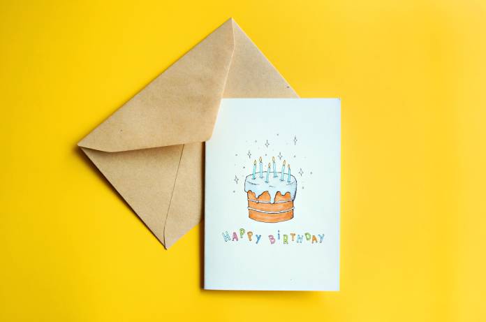 A birthday card with a hand drawn cake sits on top of a brown envelope against a bright yellow background.