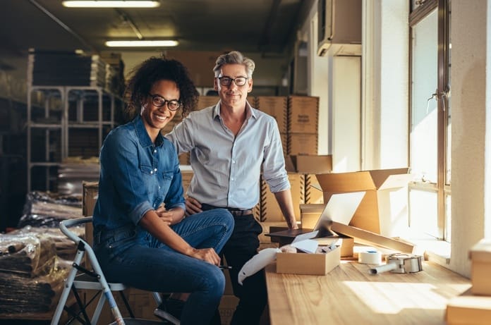 A man and a woman sitting in front of a table with packing materials on it as they run their small business.