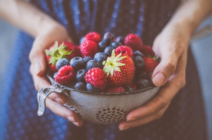 A person in a blue dress holding onto a bowl of fresh berries, including blueberries, strawberries, and raspberries.