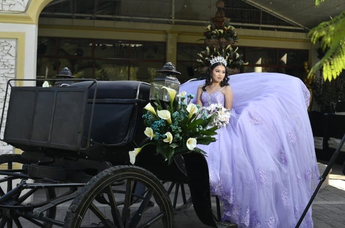 A teenage girl in a tiara and lavender ballgown with lace applique holds a bouquet of lilies as she sits in a black carriage.