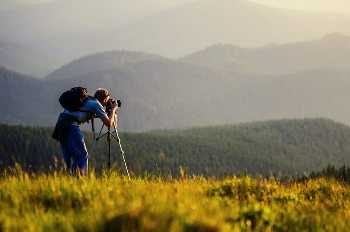 A person is standing in a field looking through a camera that sits on a tripod. Mountains appear in the background.