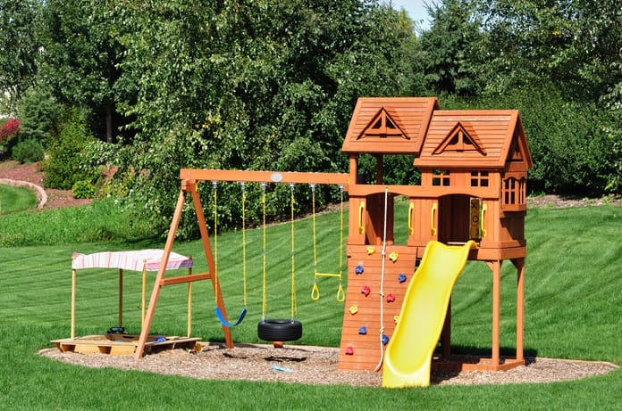 A home swing set in the backyard with a yellow slide and a wooden frame. There's mulch underneath the set.