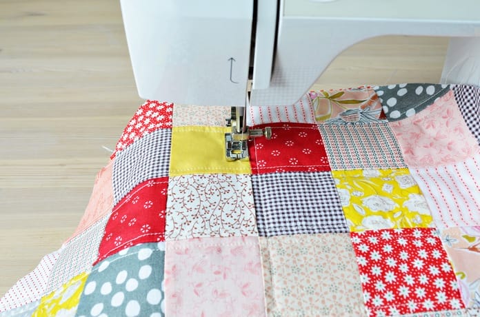 A wooden desk with a white sewing machine and a piece of patchwork fabric going through the needle head.