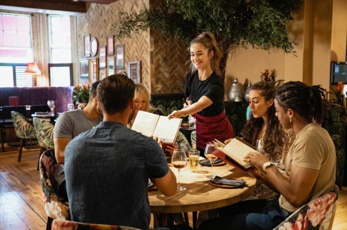 A young female waitress handing a drink menu to the table. There are five people seated at the round table.