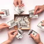 An overhead view of multiple people's hands holding one hundred-dollar bills near a donation box. The box has several bills.