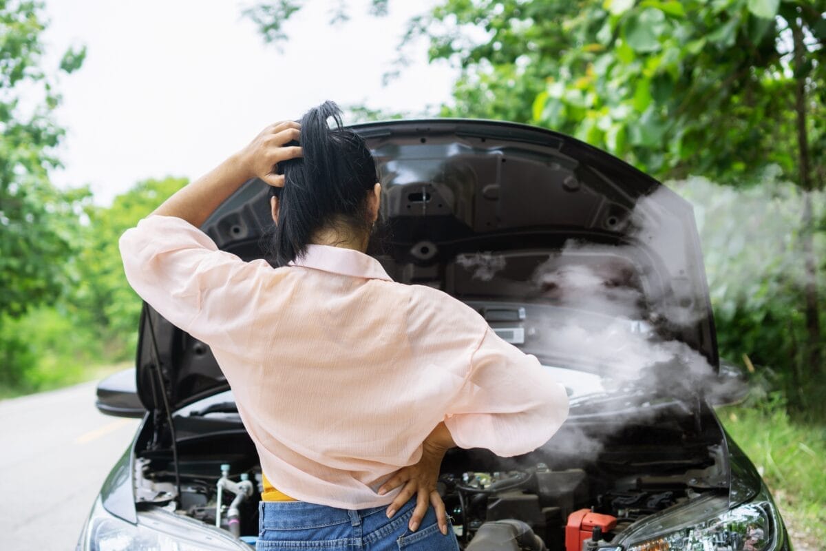 A woman stands in front of a parked vehicle with its hood open, and smoke is coming from it. She has one hand on her head.