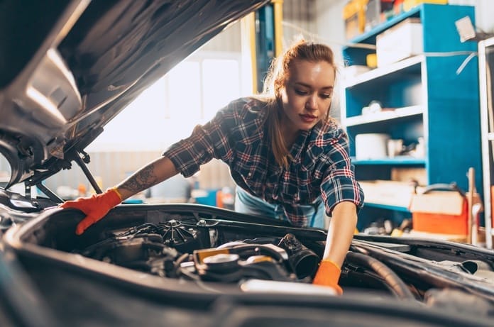 A woman working underneath the hood of a car and performing repairs. She wears orange gloves on her hands.