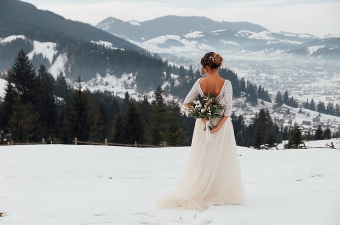 A winter bride standing in the snow in her gown and bouquet. She's facing the mountains with her bouquet behind her back.