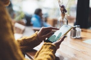 Home 18 A smartphone user holding their phone, wearing a mustard-colored sweater at a cafe table. They're typing on their phone.
