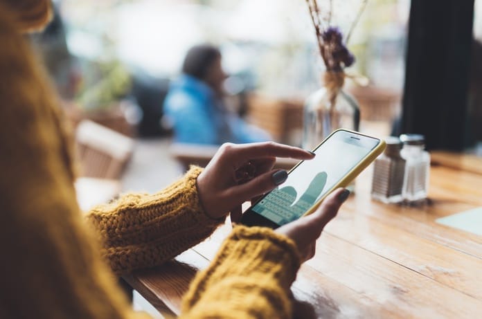 A smartphone user holding their phone, wearing a mustard-colored sweater at a cafe table. They're typing on their phone.