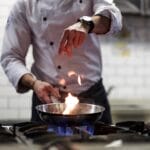 A person wearing a white chef's coat is sprinkling an ingredient into a steel pan that contains a small fire.