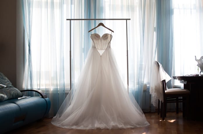 A white wedding dress with a full tulle skirt hangs on a wooden hanger in front of a large window.