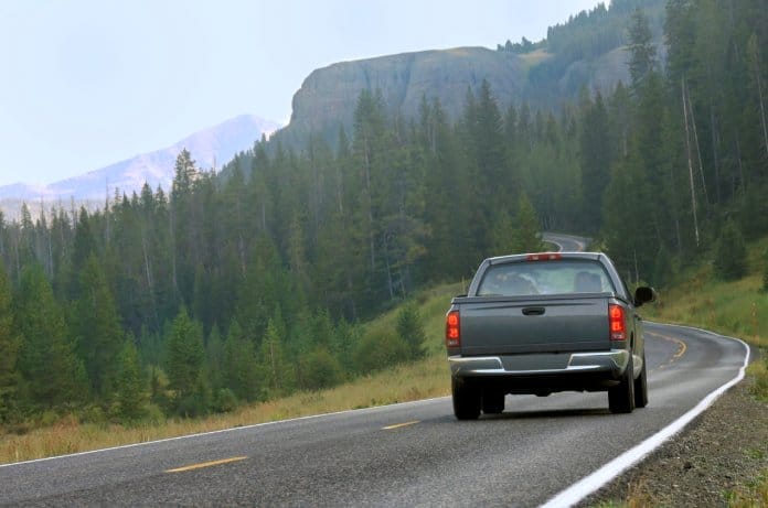 A gray pickup truck driving along a curved mountain road. Numerous trees and a cliffside are farther down the road.