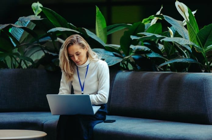 A woman in a white sweater with a blue lanyard sits on a couch in front of plants and types on a laptop.