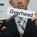A man in a suit and tie holds a pair of black scissors up to a piece of paper that says “overhead” on it.