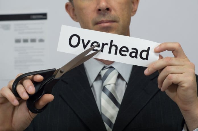 A man in a suit and tie holds a pair of black scissors up to a piece of paper that says “overhead” on it.