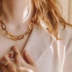 A woman in a white blazer wears a gold chain necklace around her neck and several gold rings on her hand.