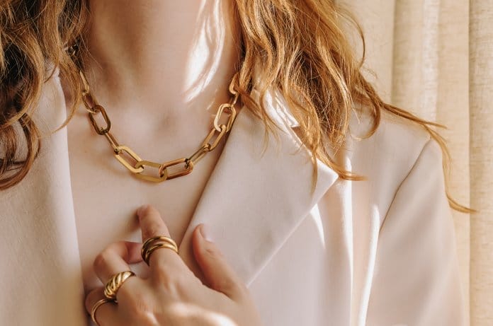 A woman in a white blazer wears a gold chain necklace around her neck and several gold rings on her hand.