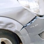 A gray car with damage to the side of the headlight and front bumper. There are scrapes on the paint and minor damage.