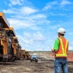A person standing in front of parked mining vehicles. He is wearing a reflective vest and a hard hat, carrying a clipboard.