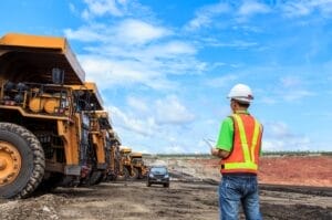 A person standing in front of parked mining vehicles. He is wearing a reflective vest and a hard hat, carrying a clipboard.
