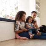 Parents and their daughter sit on the floor reading a book. They're all smiling as their daughter points at the picture.