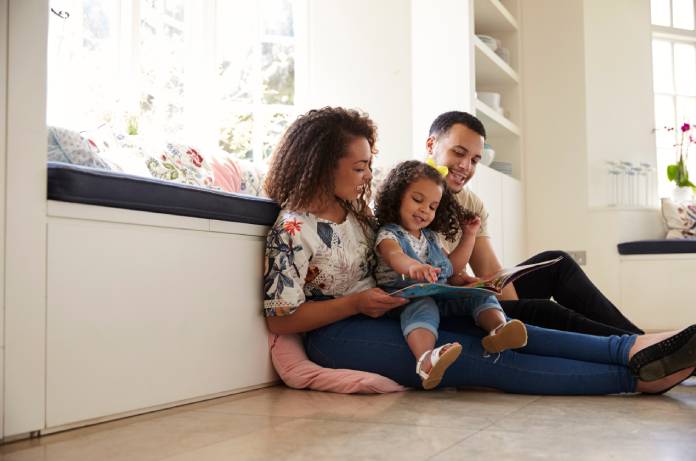 Parents and their daughter sit on the floor reading a book. They're all smiling as their daughter points at the picture.