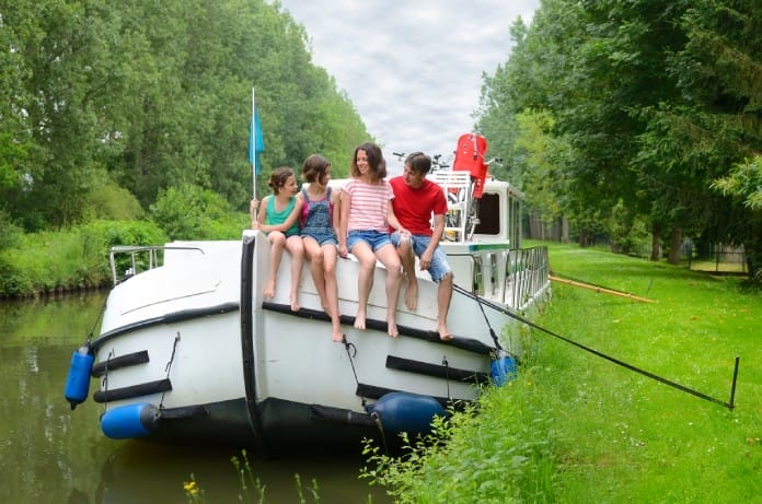 A group of kids sitting at the front of a boat on a canal that’s anchored with a rope and a stake in the ground.
