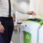 A man in professional attire holds an empty disposable plastic water bottle over a recycling bin in an office.