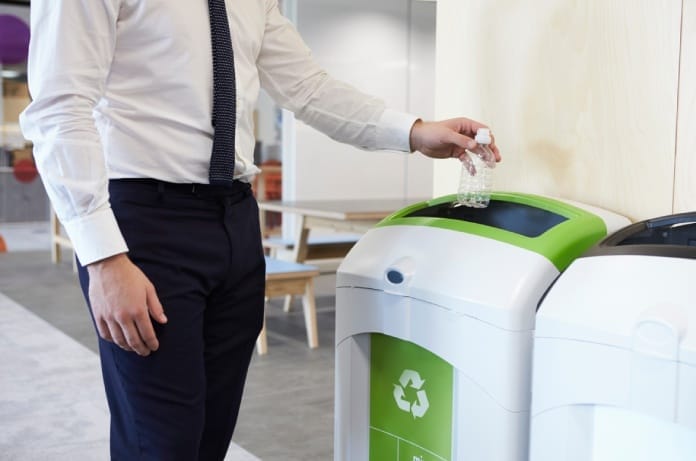 A man in professional attire holds an empty disposable plastic water bottle over a recycling bin in an office.