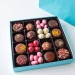 A assortment of gourmet chocolate candies are neatly arranged in a square blue box that sits on a white surface.