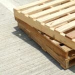 Three wooden pallets are stacked on top of each other, sitting on a concrete surface during a sunny day.
