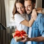 A young man and woman are hugging and smiling while the man holds a gift wrapped in brown wrapping paper and a red bow.