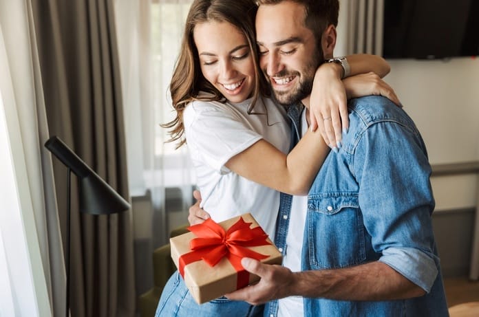 A young man and woman are hugging and smiling while the man holds a gift wrapped in brown wrapping paper and a red bow.